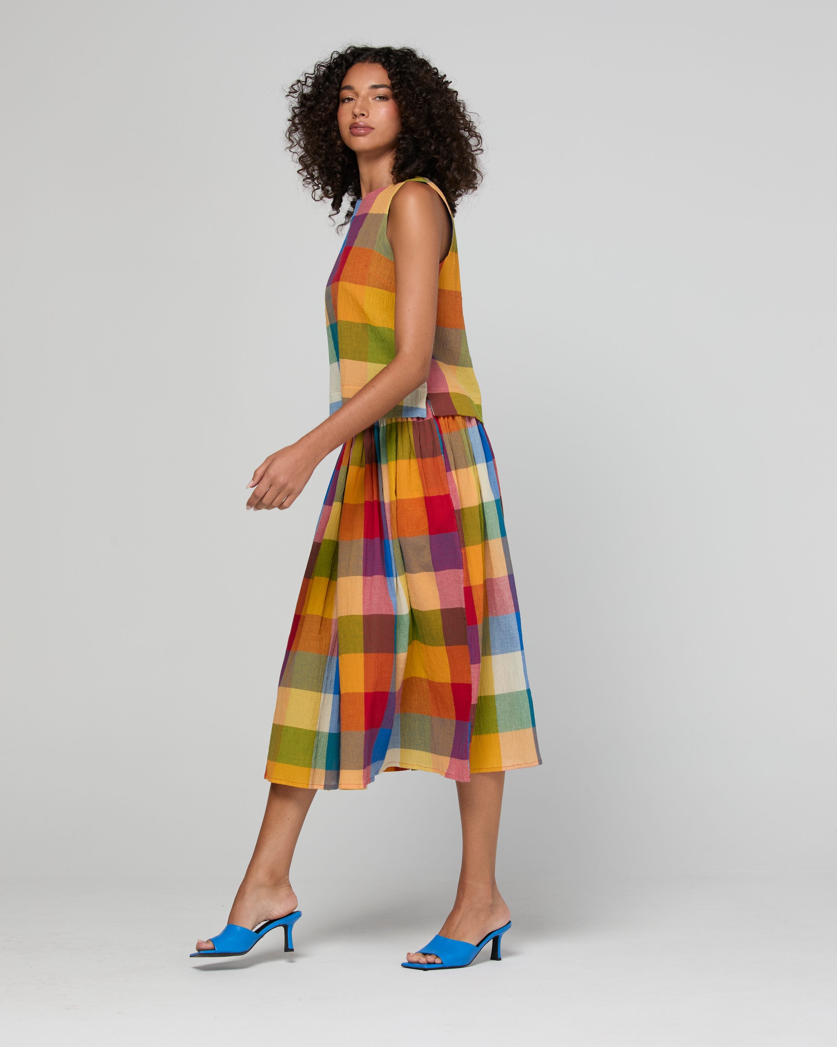 A woman modeling a sleeveless top and mid length skirt with a colorful plaid pattern in shades of orange, yellow, green, red, and blue, paired with bright blue heeled mule sandals, against a light gray studio background.