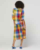 A model is shown from the back wearing a colorful plaid shirt with three-quarter puffed sleeves and blue heeled mules, set against a plain light gray studio background.