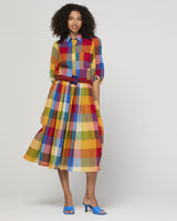 A woman modeling a colorful plaid shirt dress with a matching belt and blue heeled sandals against a light gray studio background.