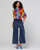A model wearing a vibrant multicolor patchwork plaid shirt with blue wide-leg cropped pants and blue heels, standing against a light gray studio background.