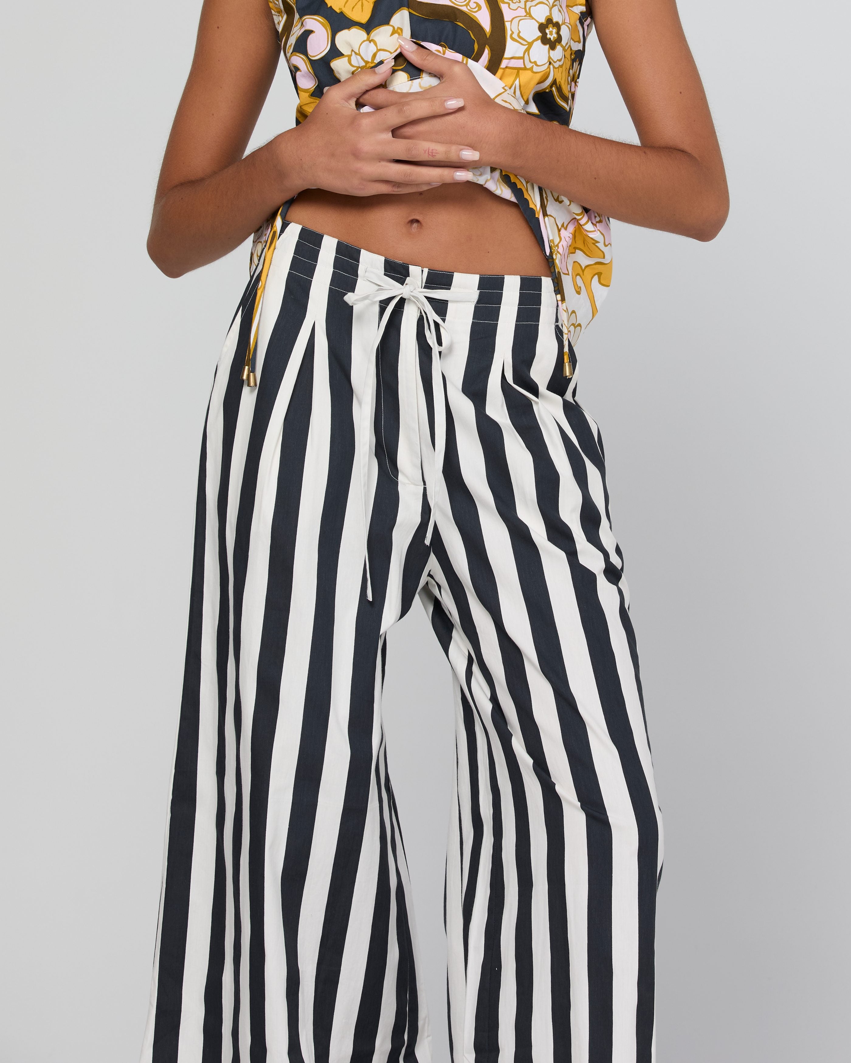 A model wearing high-waisted, wide-leg pants with bold vertical black and white stripes, paired with a patterned crop top, against a neutral gray background.