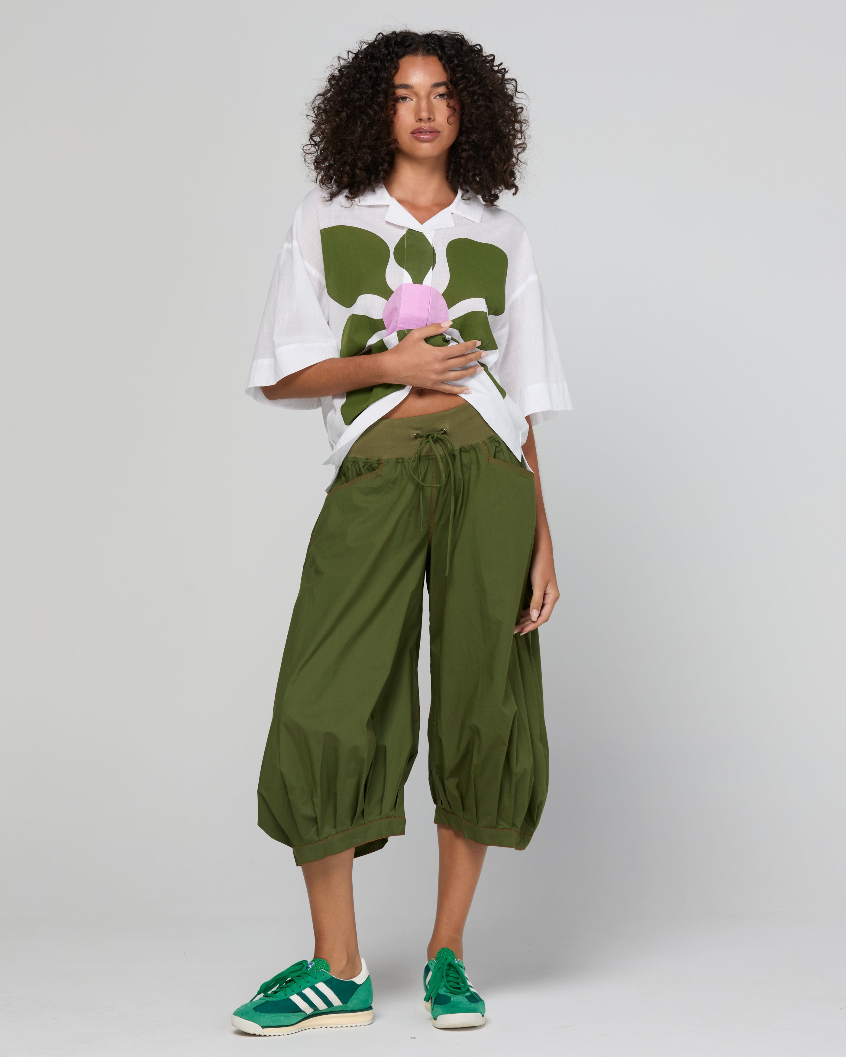 Woman wearing a white shirt with a floral design and green pants on a plain background