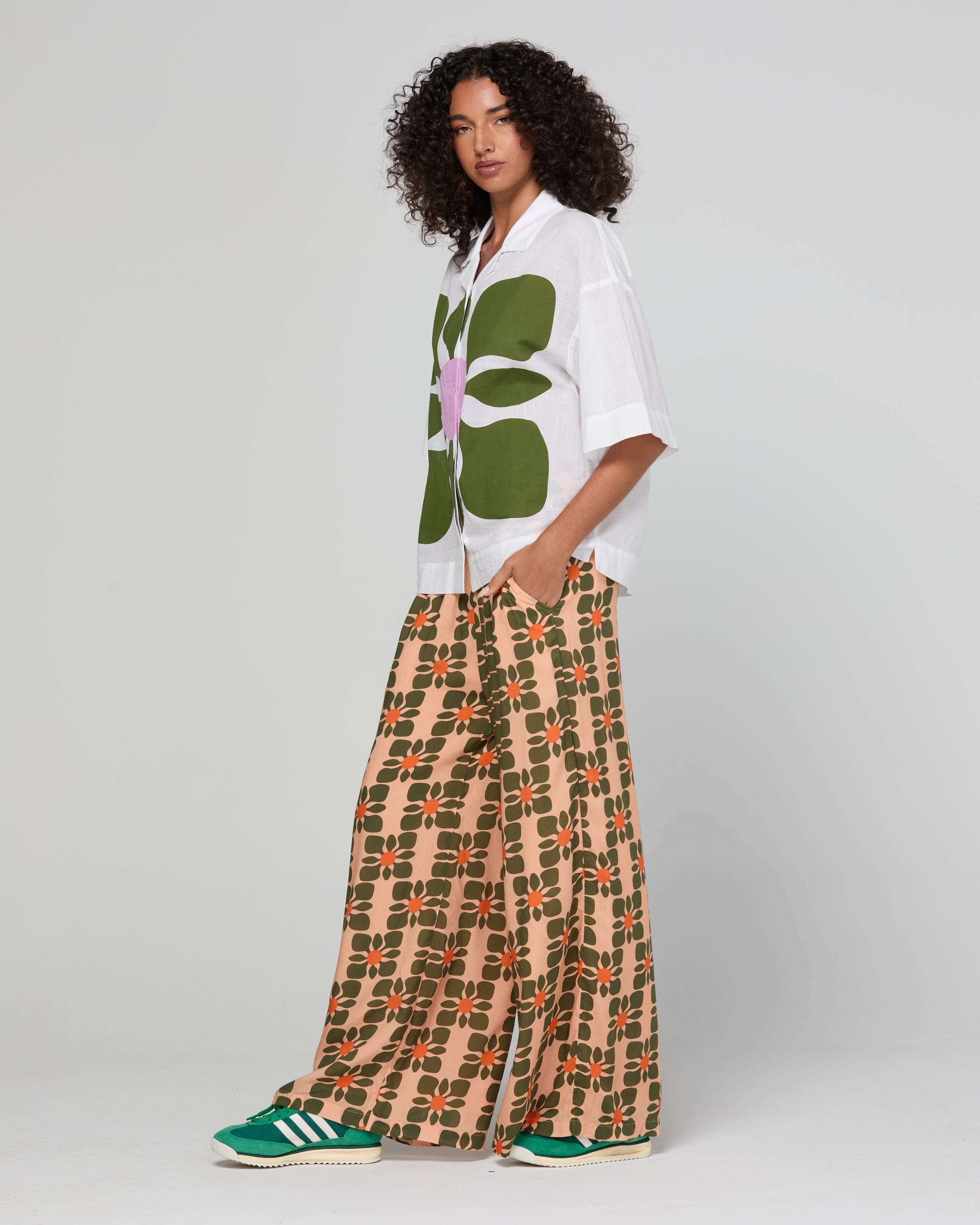 Woman wearing a white shirt with green floral design and patterned beige pants on a gray background