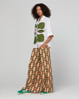 Woman wearing a white shirt with green floral design and patterned beige pants on a gray background