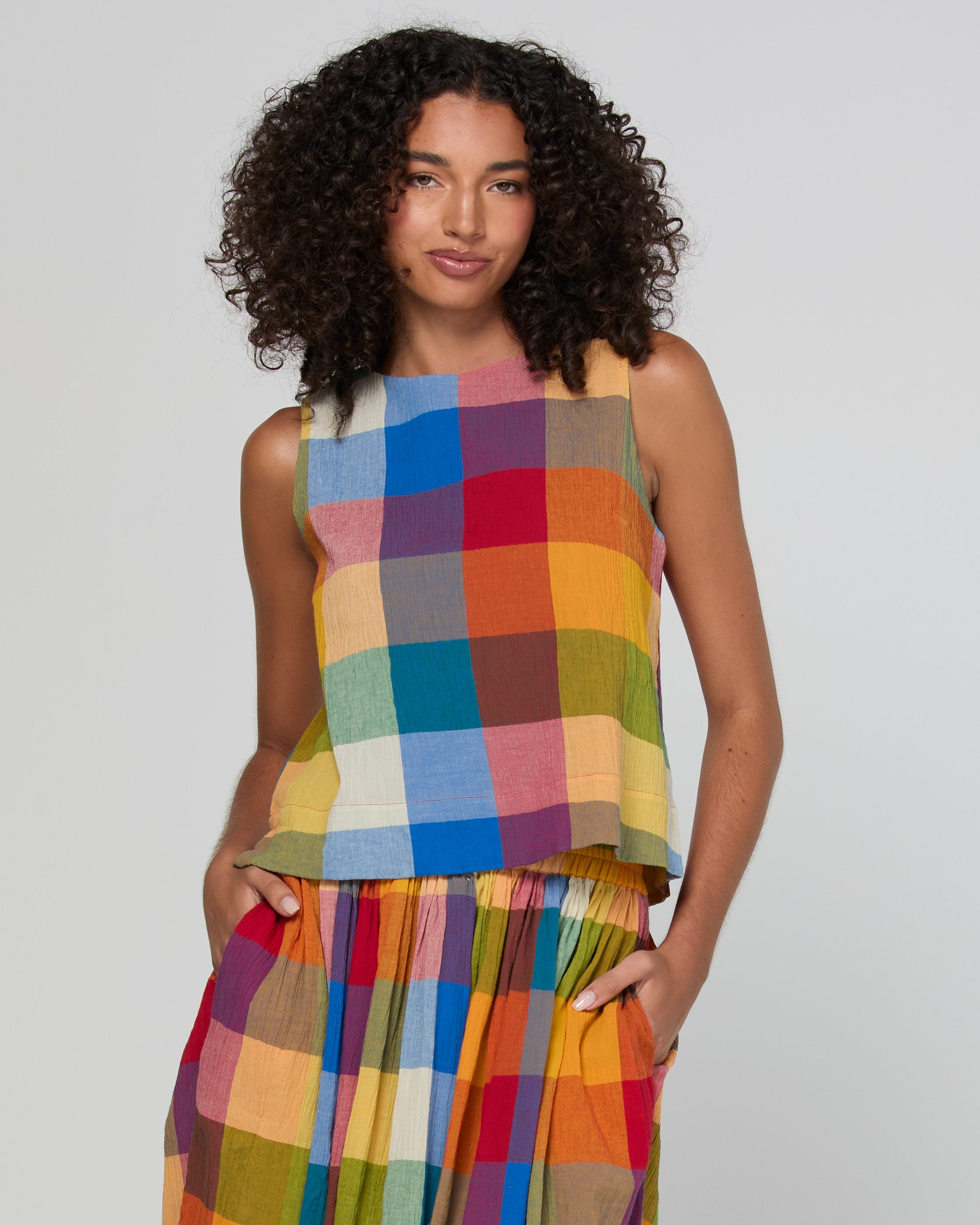 A model with curly dark hair wearing a sleeveless top and matching pleated skirt set in a vibrant multicolor plaid pattern, set against a plain light gray studio background.