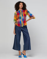 A model stands against a light gray studio background wearing a colorful patchwork button-up shirt with three-quarter sleeves, dark blue wide-leg cropped pants, and bright blue heeled sandals.
