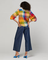 A model is shown from behind wearing a colorful plaid puff-sleeve blouse and dark blue wide-leg cropped pants, paired with blue mule heels, against a light gray studio background.