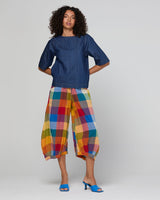A model stands against a light gray studio background wearing a dark blue short-sleeve top and colorful plaid cropped pants in shades of yellow, red, blue, green, and orange, paired with blue heeled sandals.