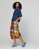 A model wearing a dark blue cropped t-shirt, colorful rainbow plaid cropped pants, and blue mule heels, photographed against a solid light gray studio background.