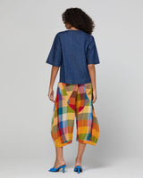 A model shown from the back wearing a dark blue short-sleeve top and vibrant, multicolored geometric plaid cropped pants, paired with blue heels, against a light gray studio background.