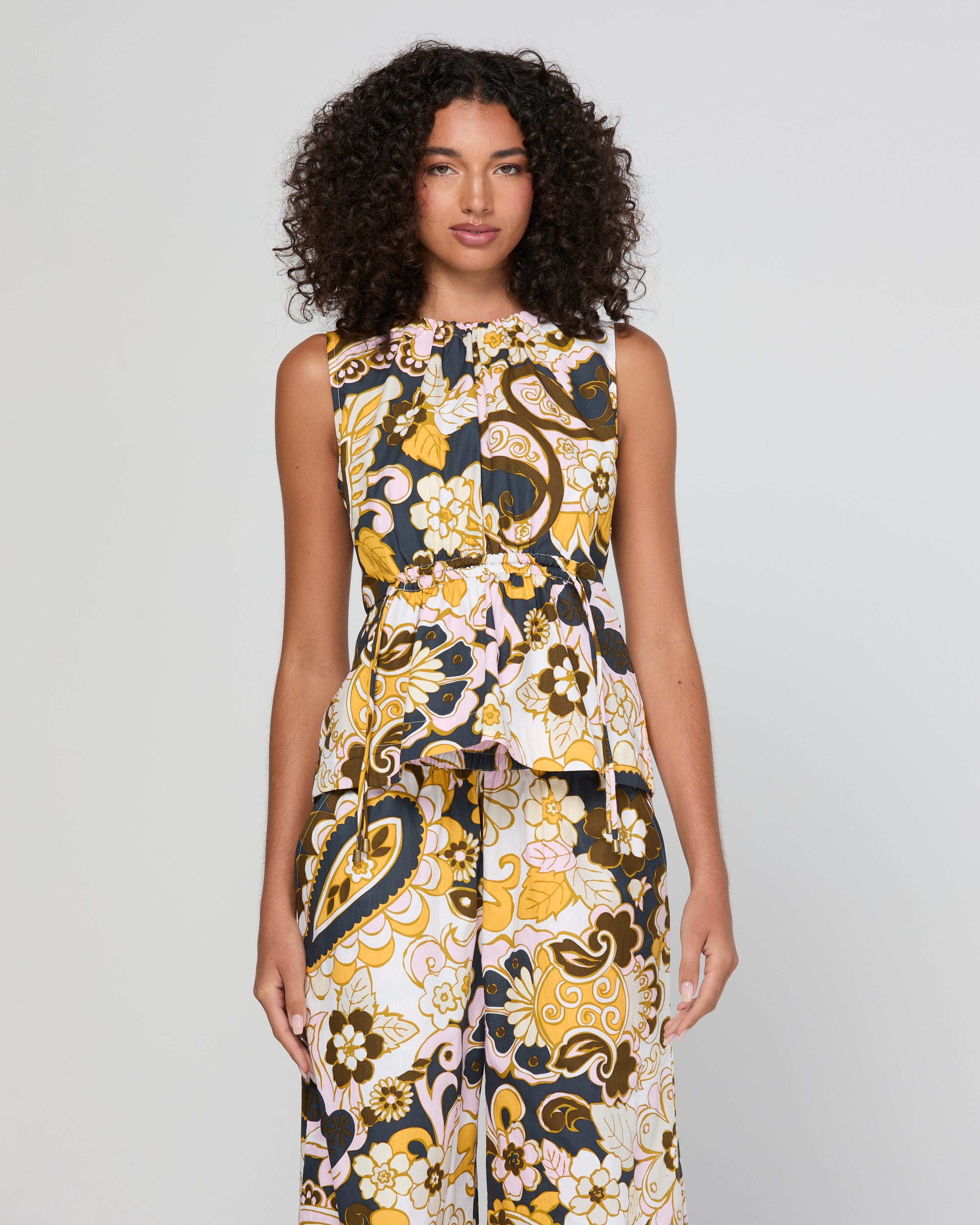A woman with curly dark hair models a sleeveless jumpsuit featuring a bold, retro paisley and floral pattern in yellow, brown, black, and white against a light gray studio background.