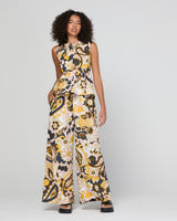 A woman modeling a sleeveless, wide-leg jumpsuit with a bold paisley pattern in black, yellow, and cream, set against a plain light gray studio background.