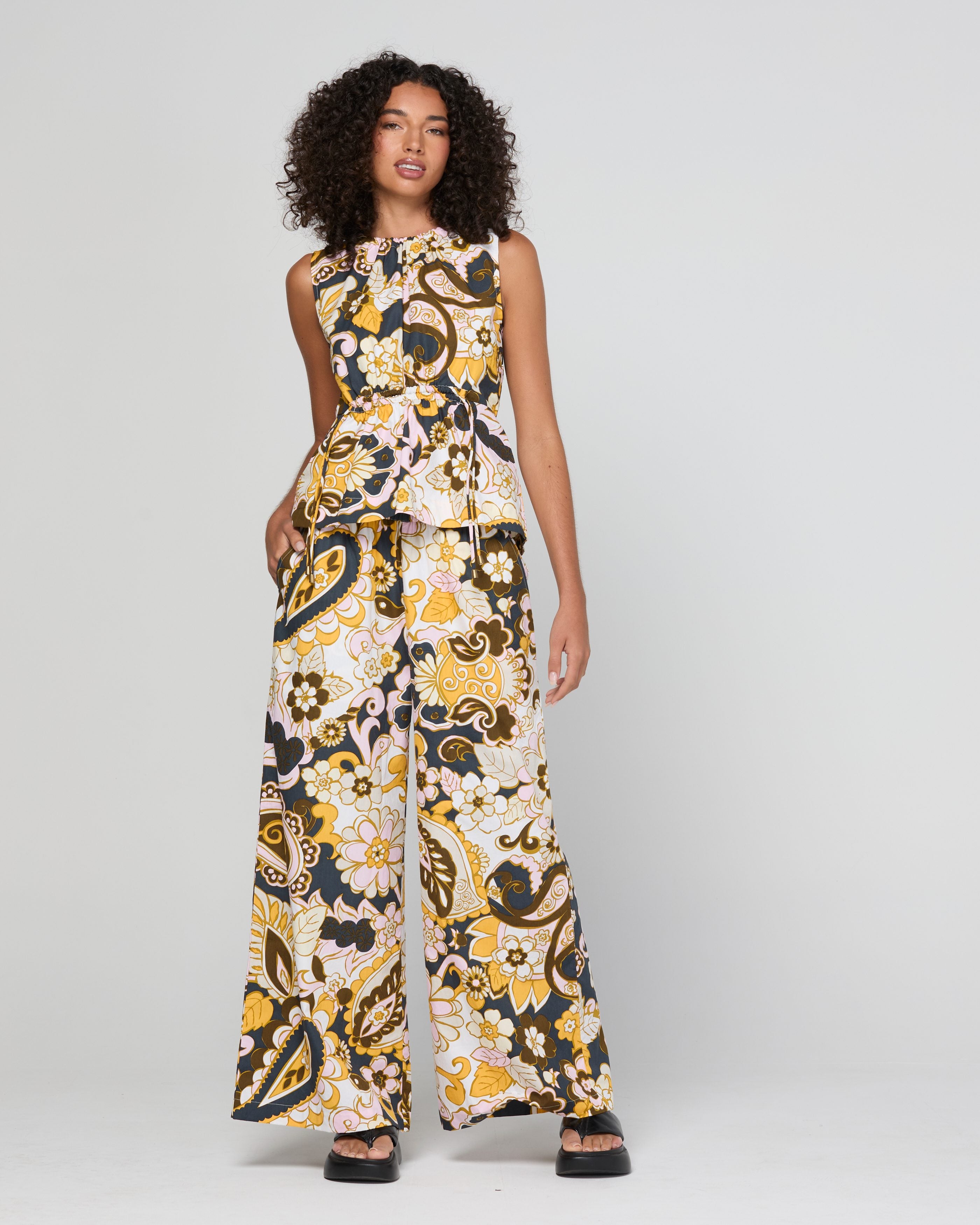 A woman modeling a top, wide-leg pant with a bold abstract pattern in black, yellow, and cream, set against a plain light gray studio background.