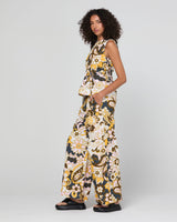 A model wearing a sleeveless matching set with a wide-leg pant and a ruffled peplum top, featuring a bold yellow, black, and white floral pattern, photographed against a light gray studio background.