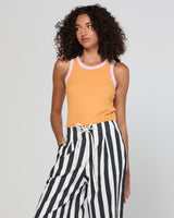 A woman modeling a sleeveless tank top in mandarin colour with light pink trim, paired with black and white vertically striped wide-leg pants, against a light gray studio background.