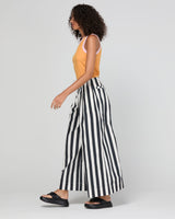 A woman modeling a sleeveless tank top in mandarin colour with light pink trim, paired with black and white vertically striped wide-leg pants, against a light gray studio background.