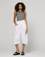 A model wearing a black and white striped sleeveless crop top, white high-waisted cropped pants, and black slide sandals, standing against a light gray studio background.
