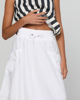 A close-up of a person wearing a white A-line skirt with a drawstring waist and side pockets, paired with a black and white striped top, against a light gray background.