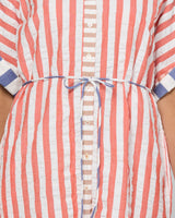 Close-up of a short-sleeve striped shirt dress with vertical coral pink and white stripes, a button front, and a thin blue tie belt at the waist.