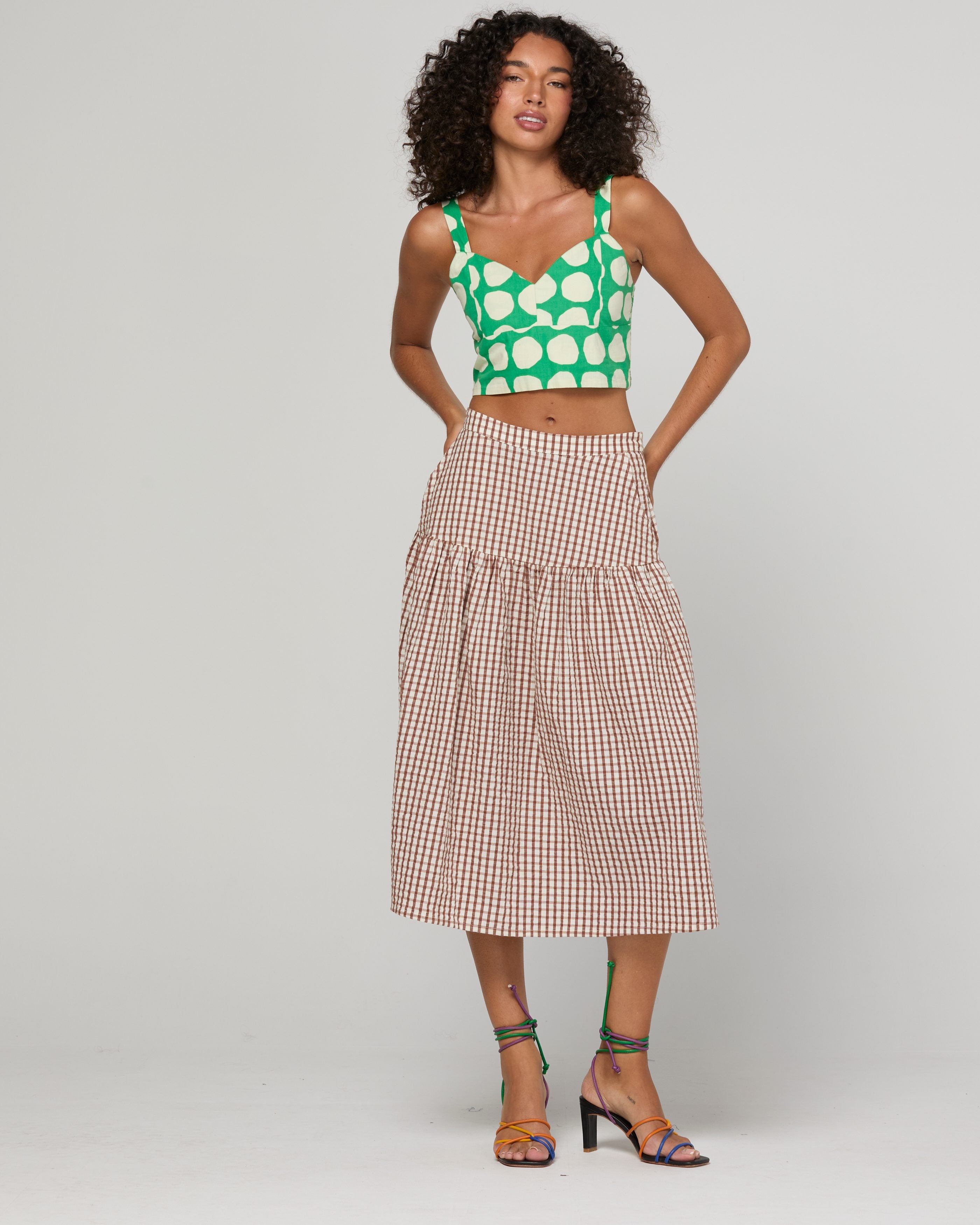 A model stands against a light gray studio background wearing a green and white polka-dot crop top with beige and white gingham tiered midi skirt and green strappy high-heeled sandals.