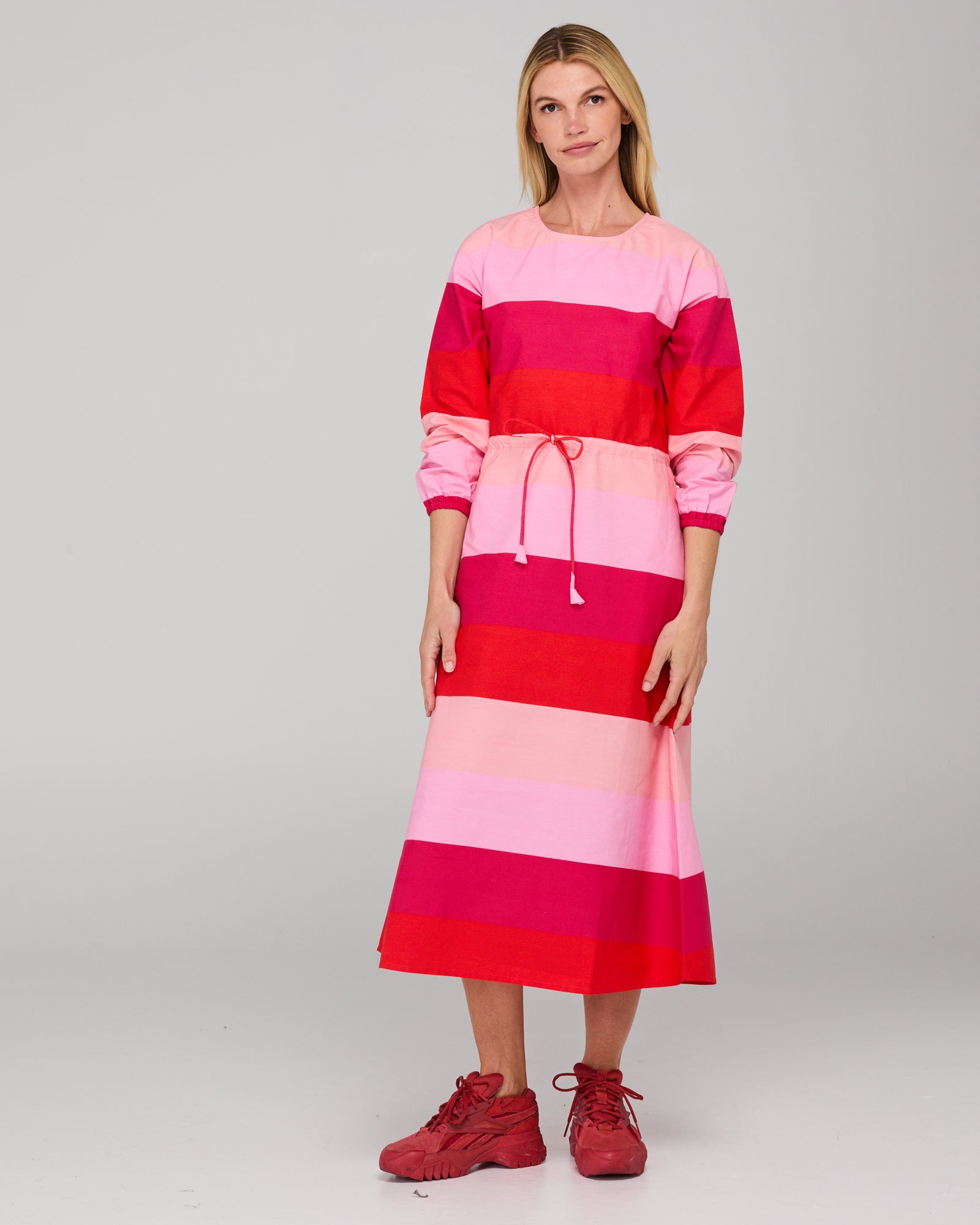 Woman wearing a long-sleeved pink and red striped midi dress on a gray background