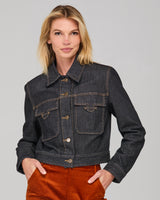 Woman wearing a dark denim jacket and rust-colored cords against a gray background