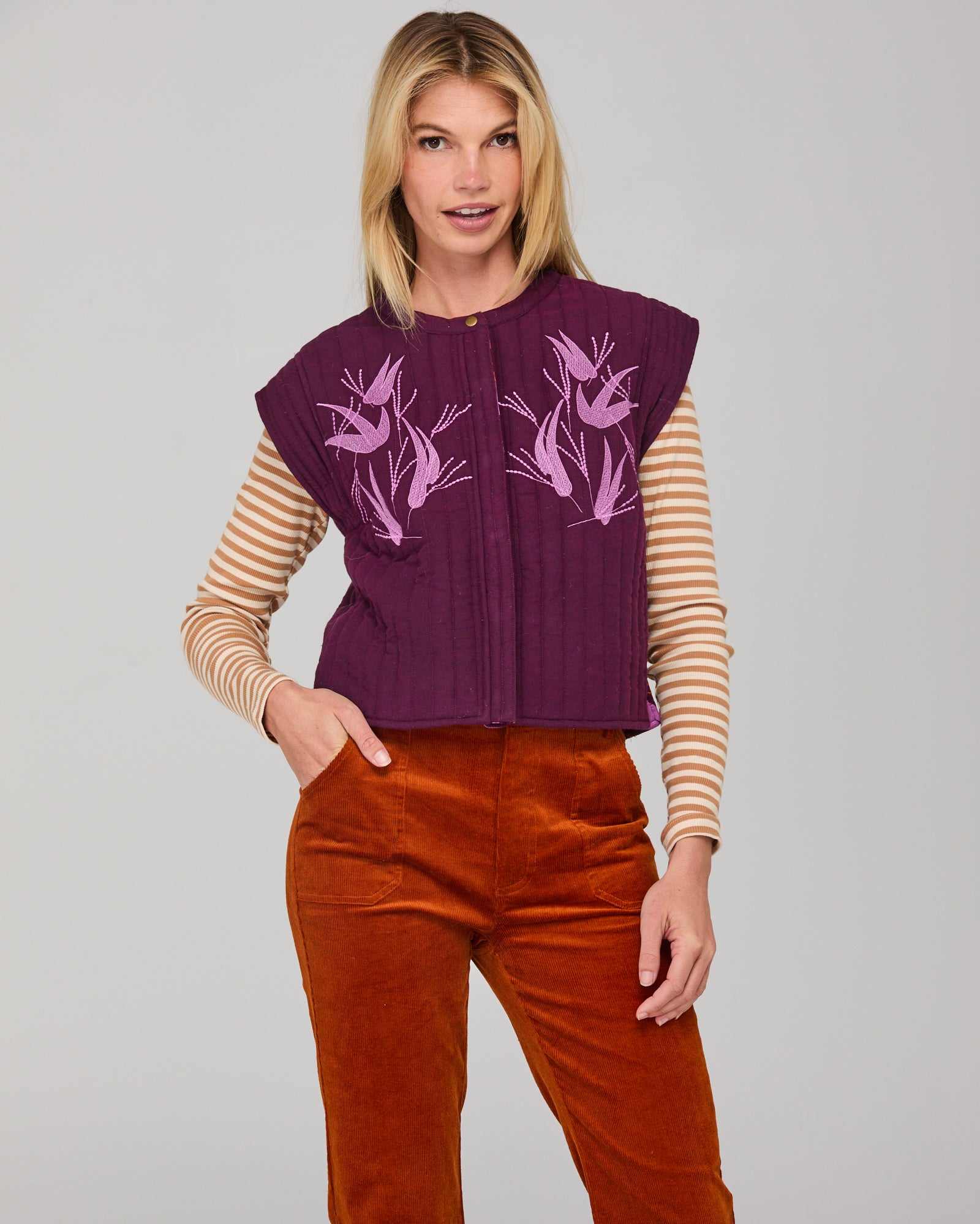Woman wearing a purple vest with floral patterns, cream striped shirt, and rust cord pants on a gray background