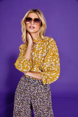 Woman wearing a yellow block print floral blouse and ditsy patterned pants against a purple background