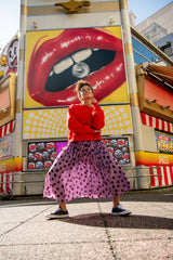 Fashion model in a red sweater and polka dot skirt standing in front of a colorful billboard with a large mouth graphic.