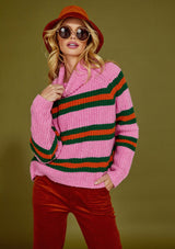 Woman wearing a pink sweater with green and orange stripes, rust pants, and a rust bucket hat against a green background.